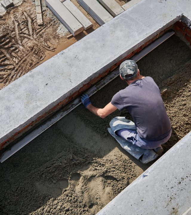 Top view of male contractor placing screed rail on the floor covered with sand-cement mix at construction site. Man worker leveling surface with straight edge while screeding floor in new building.